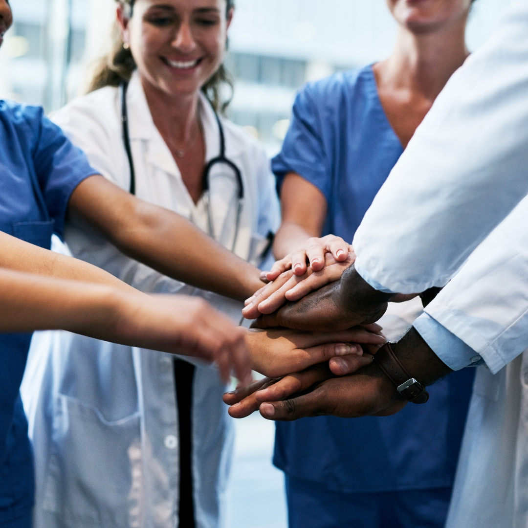 Grupa pracowników medycznych w fartuchach i uniformach pielęgniarskich kładzie dłonie jedna na drugiej w geście współpracy i jedności. Widać uśmiechy i pozytywną atmosferę, symbolizującą pracę zespołową w ochronie zdrowia.